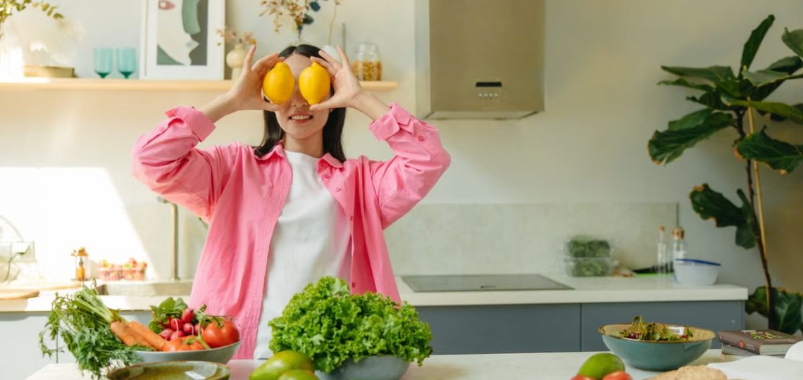 A woman playfully holding lemons over her eyes in a bright, lively kitchen setting filled with fresh vegetables.